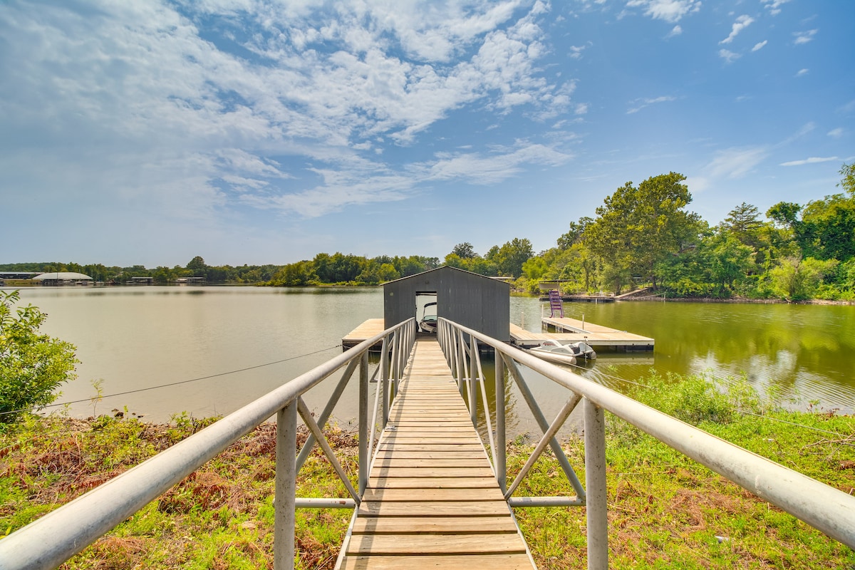 A wooden dock extends toward the water, framed by lush greenery. On either side, grassy areas lead to the lake's edge, with gentle waves reflecting the clear sky. Structures for boat docking are visible at the water's end.
