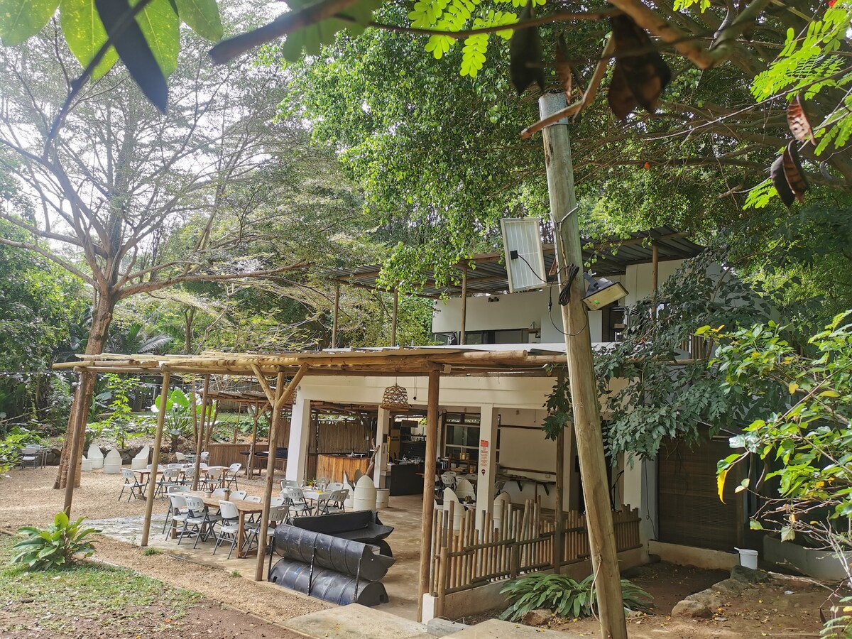 A two-story building is surrounded by lush greenery, featuring an open terrace that overlooks the area. The ground level includes a dining space with multiple tables and chairs, while the upper level provides a shaded area with seating options for relaxation amidst nature.