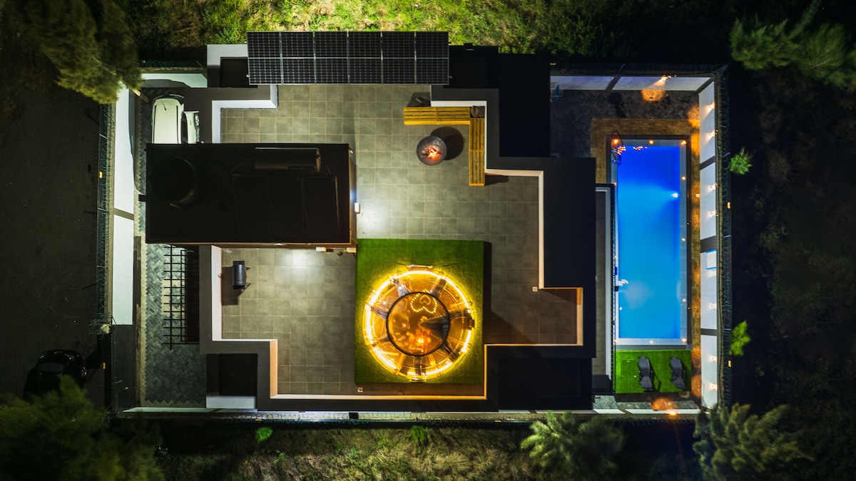 The aerial view captures the villa's layout, showcasing the private swimming pool surrounded by sunbeds and a central fire pit area elegantly illuminated. Solar panels are visible on the roof, highlighting the eco-friendly design of the property.