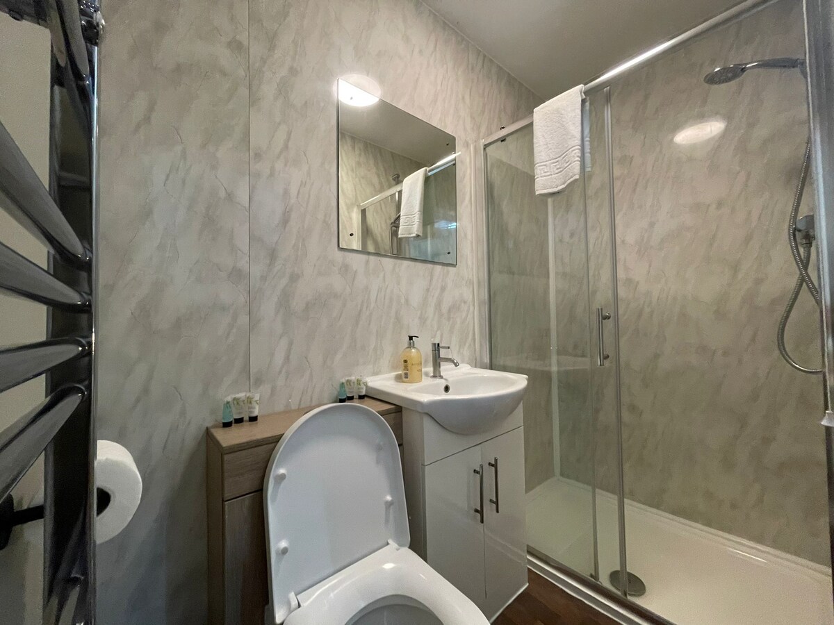 An ensuite bathroom showcases a modern design with a refreshing shower, a built-in sink, and a toilet. A full-length mirror is mounted above the sink, while complimentary toiletries and fresh towels are neatly arranged. Soft natural light enhances the serene atmosphere.