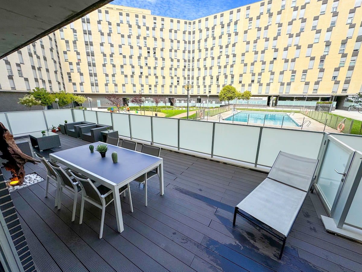 A spacious terrace features a large dining table surrounded by comfortable chairs, ideal for outdoor meals. A single lounger is positioned nearby, while a well-maintained pool area is visible in the background. Low-lying plants add a touch of greenery to the space.