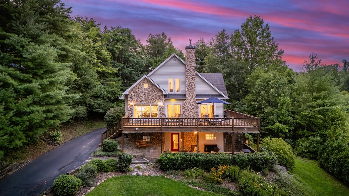 The exterior of the house is surrounded by lush greenery and features stone and wood elements. A spacious deck wraps around the front, providing outdoor seating. The warm glow of lights emanates from the windows, contrasted by a vibrant sunset sky in shades of pink and purple.