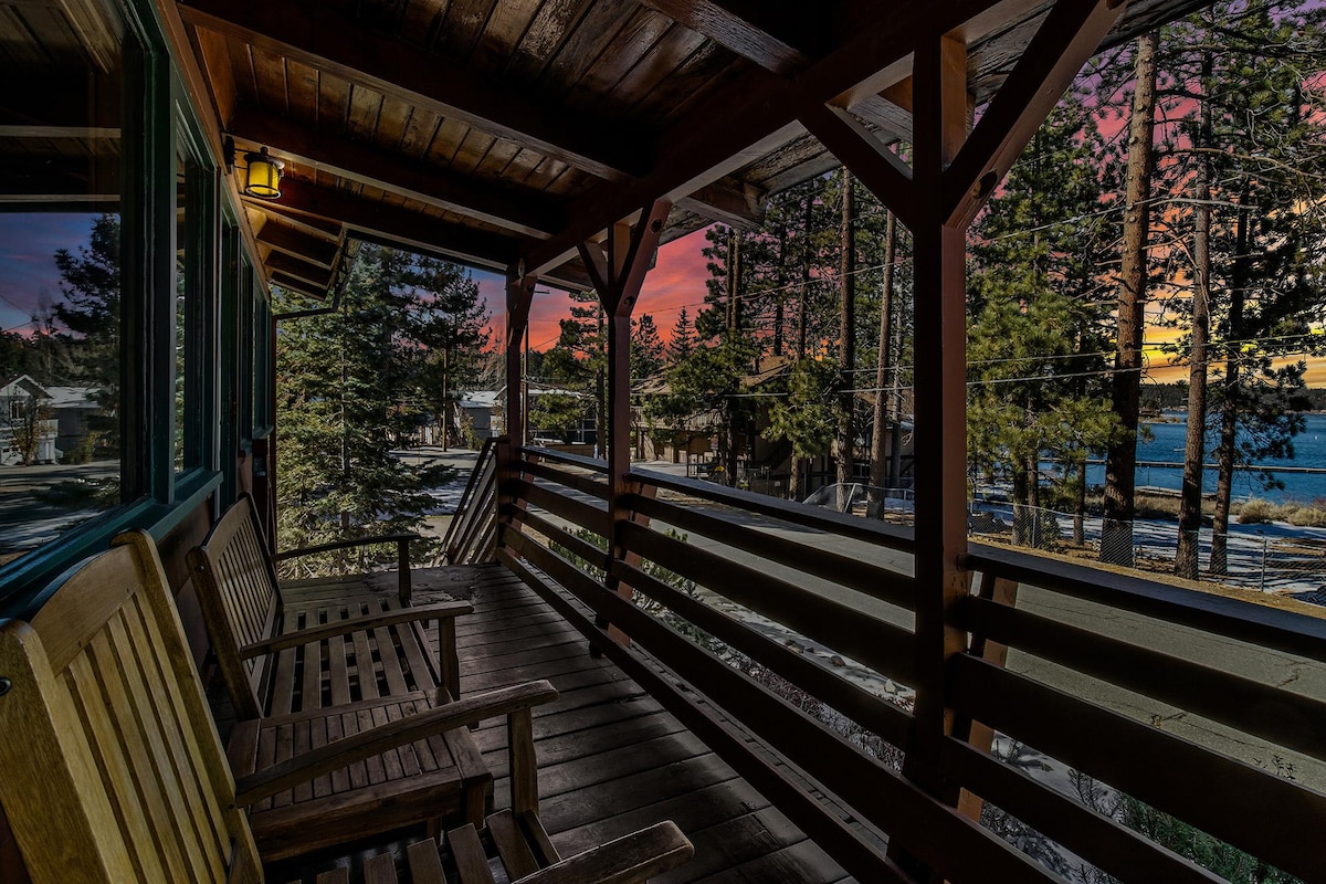 A covered front porch features wooden seating facing a serene landscape. Tall trees are visible, with a glimpse of Big Bear Lake in the distance. The sky is painted with vibrant colors, indicating a sunset, creating a peaceful outdoor setting.