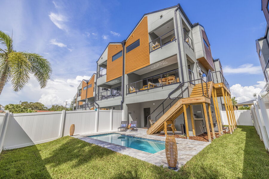 The exterior of the three-story townhome is depicted, showcasing a modern design with a combination of gray and orange accents. A private pool is situated in the backyard, framed by manicured grass, with steps leading up to the entrance of the home.