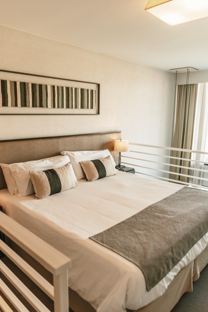 A well-appointed bedroom features a spacious bed adorned with neutral bedding and accent pillows. Soft natural light filters through sheer curtains, and a piece of abstract wall art complements the decor. A bedside lamp adds functionality to the inviting space.