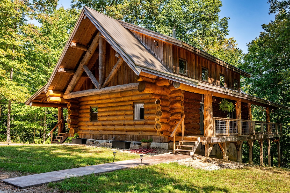 A rustic log cabin with a large sloped roof is set among lush green trees. The exterior features natural wood logs and large windows, creating a warm and inviting appearance. A concrete path leads to the entrance, surrounded by an expansive lawn.