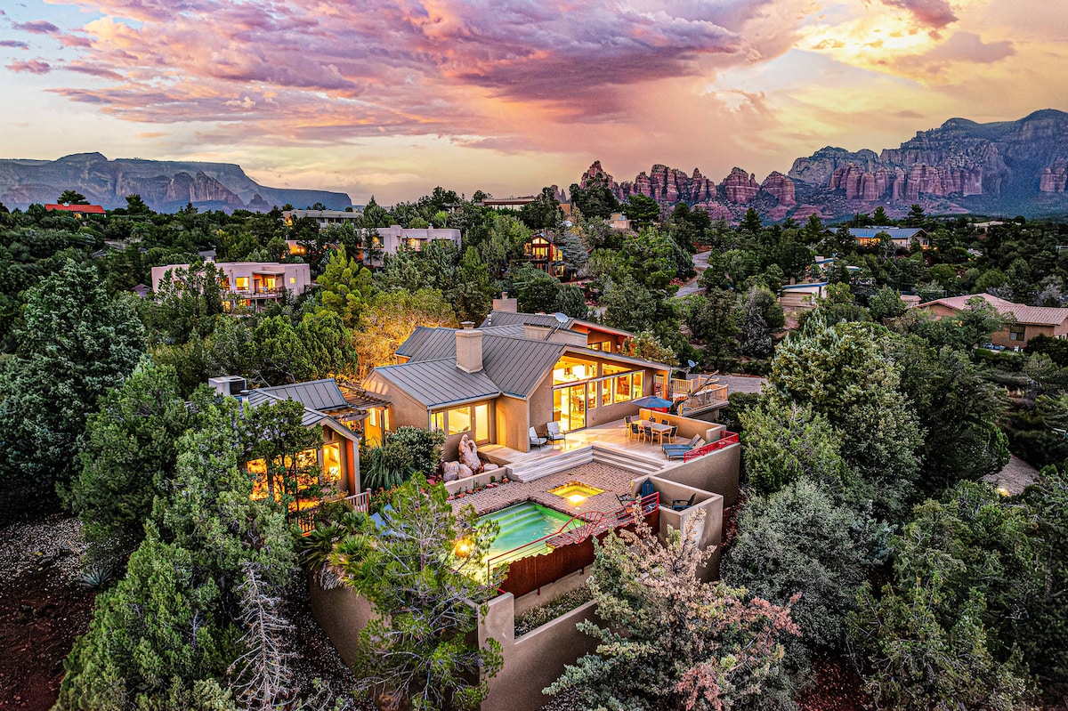 An aerial view showcases a modern home surrounded by lush greenery, featuring a salt-water pool and expansive deck. Colorful clouds are visible against the backdrop of Sedona's iconic rock formations, creating a serene landscape. The architecture blends harmoniously with the natural setting.