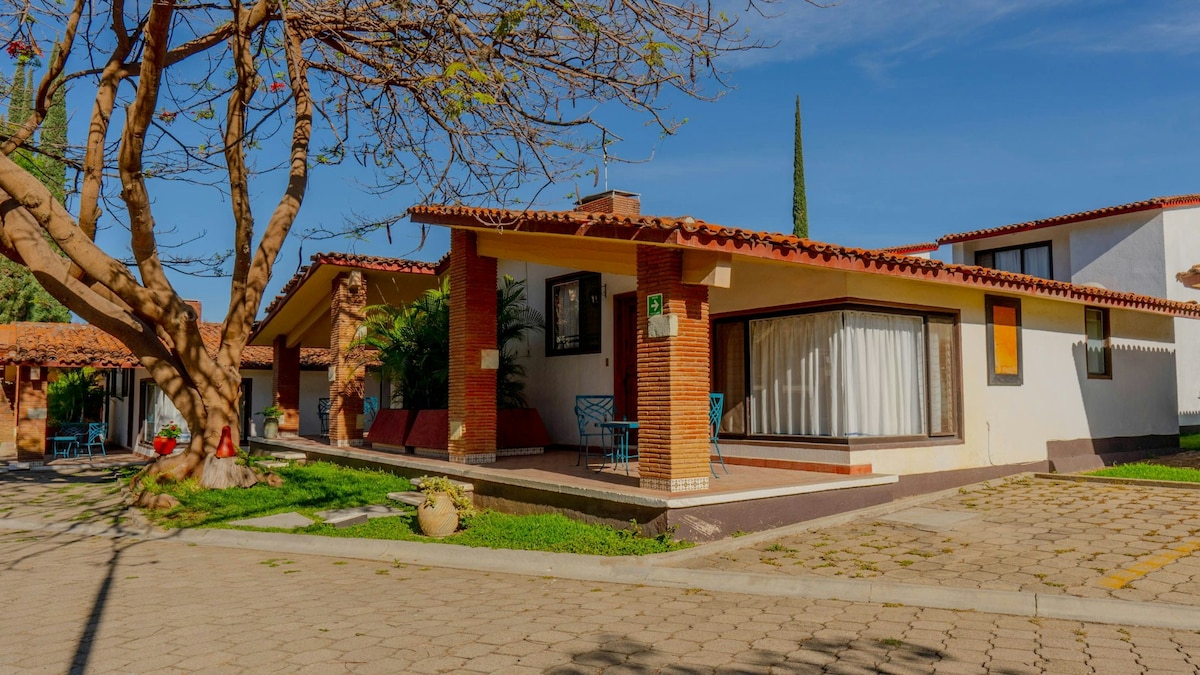 A charming villa features a welcoming porch with bright blue chairs, surrounded by lush greenery. The building presents a combination of stone and white walls under a red-tiled roof, alongside a well-kept pathway and trees that enhance the outdoor space.