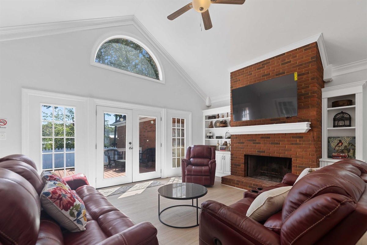 A spacious living area features leather reclining chairs and a circular glass coffee table. A large window allows natural light, while a brick fireplace serves as the focal point. French doors lead to an outdoor space, enhancing the room's open feel.