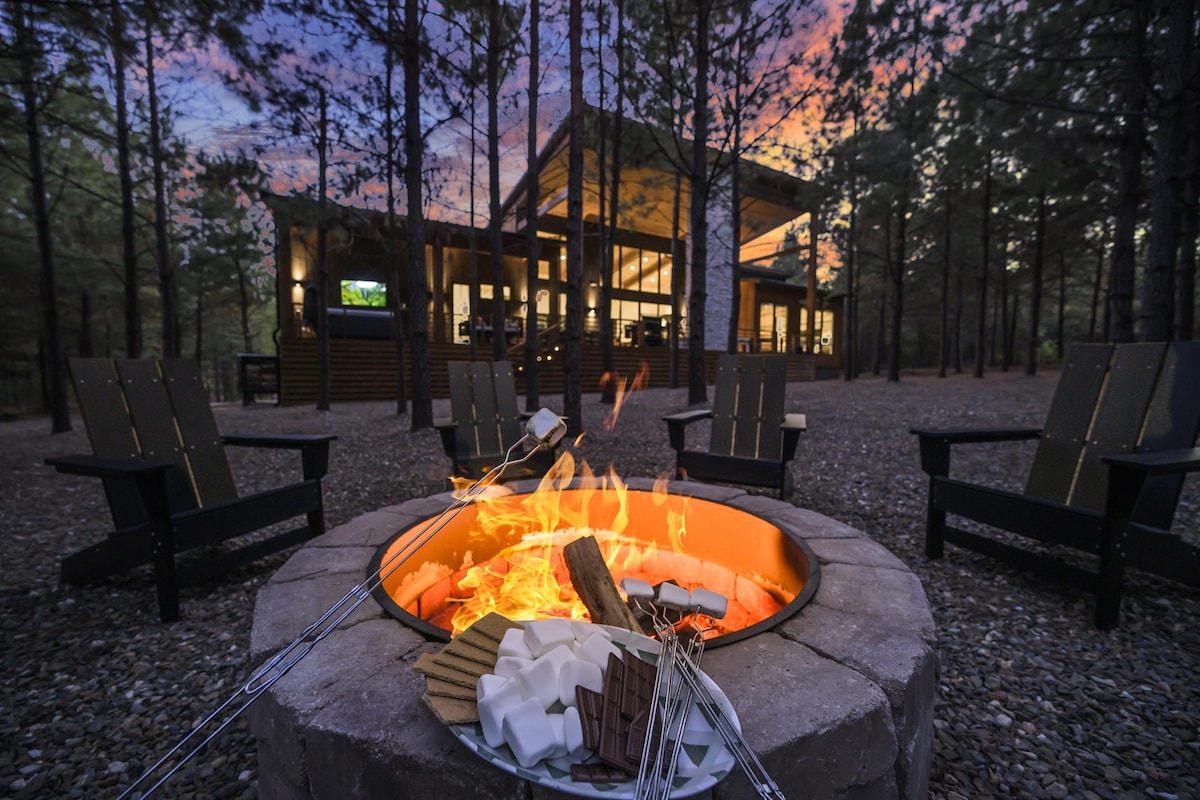 An inviting fire pit is surrounded by multiple wooden chairs, creating a cozy gathering spot. S'mores ingredients are arranged on a plate beside the fire. The background showcases the cabin, framed by tall pine trees and a colorful sunset sky.