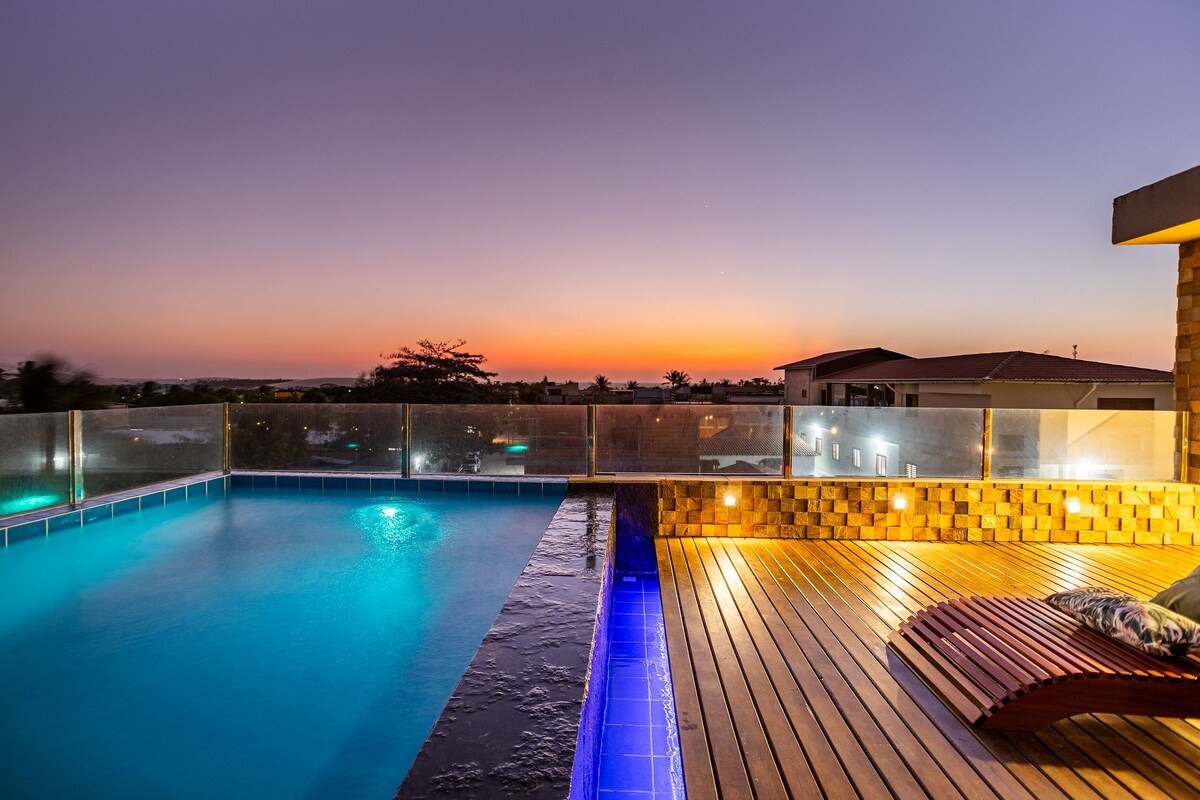 A tranquil rooftop pool is illuminated by warm lights, providing a contrast to the vibrant sunset sky. The pool's edge seamlessly meets a wooden deck, enhancing the serene ambiance. The view beyond captures the landscape and distant buildings, creating an inviting space for relaxation.