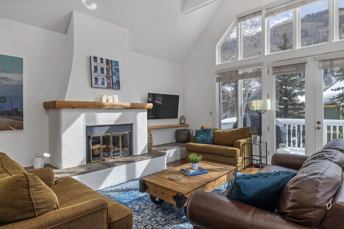 A spacious living area features a large gas fireplace framed by a wooden mantle. Soft brown sofas are arranged around a rustic coffee table, with a blue area rug beneath. Large windows allow natural light and mountain views, creating a bright and inviting atmosphere.