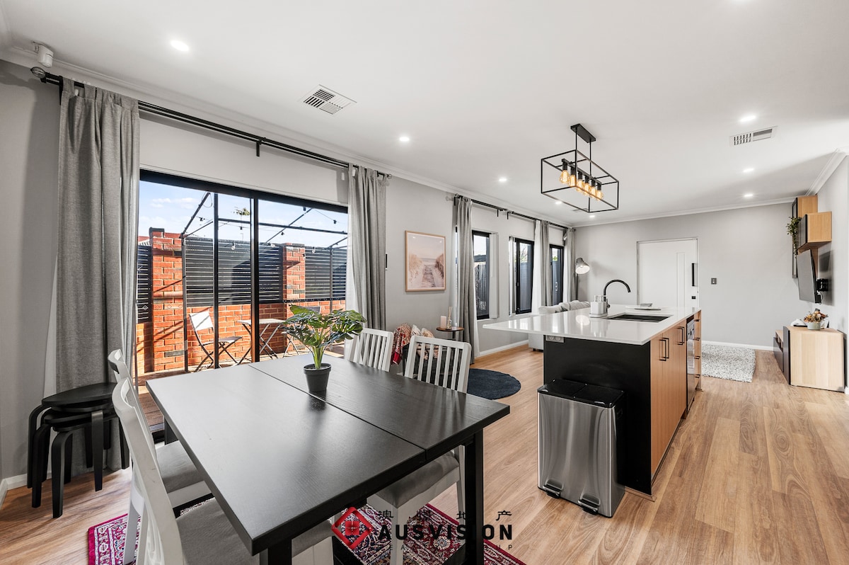 The modern open-plan kitchen and dining area showcases a large island, equipped with quality appliances. A dining table is surrounded by colorful chairs, while large windows invite natural light. The space features neutral tones and stylish décor, enhancing the inviting environment for gatherings.