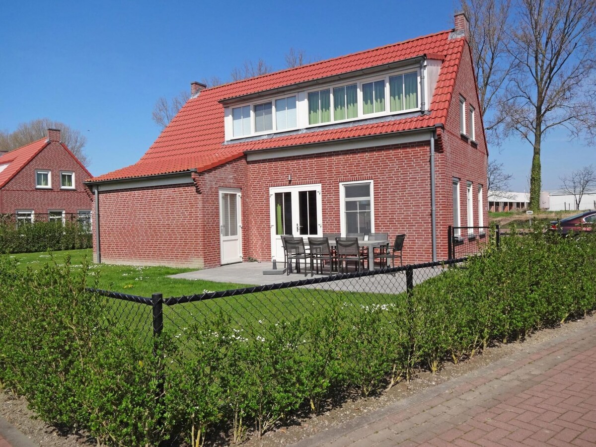 A two-story house featuring a red-brick exterior and a red tiled roof. Large windows enhance the bright appearance. A spacious patio area with outdoor furniture is visible, surrounded by a neatly maintained garden enclosed by a fence.
