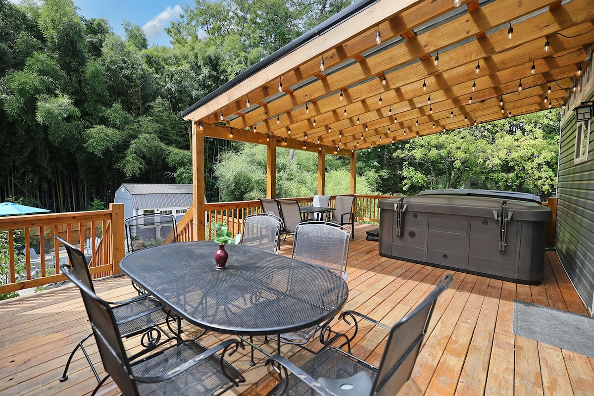 A spacious outdoor deck is highlighted by a large table surrounded by six metal chairs. A hot tub is positioned nearby under a wooden pergola, with string lights adding soft illumination. Lush greenery and a distant structure create a serene atmosphere.