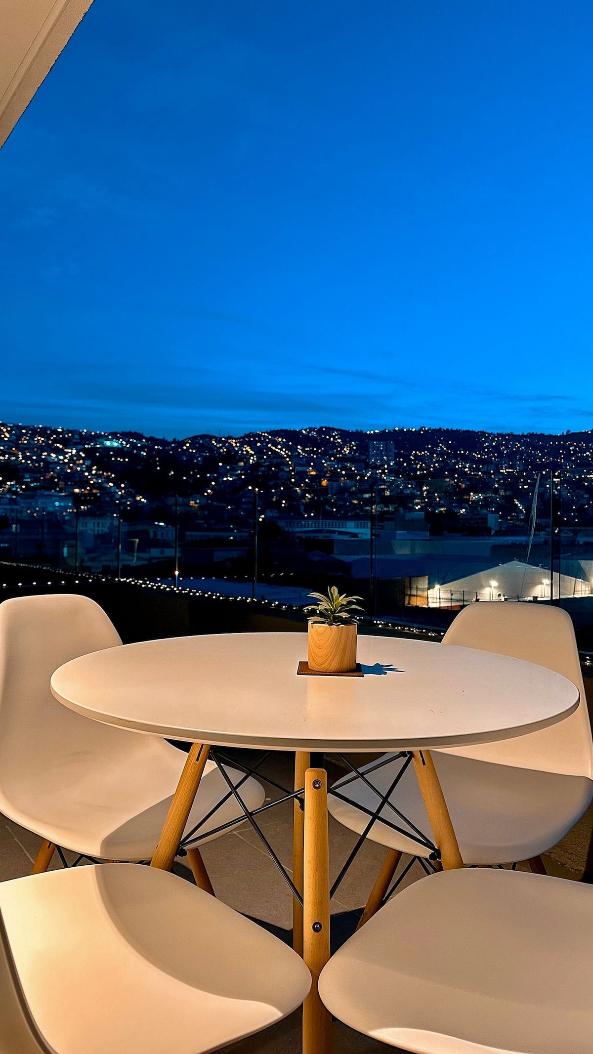 A round table is set on a balcony, accompanied by four white chairs. A small potted plant sits in the center of the table. The distant hills are illuminated by city lights under a deep blue sky, creating a serene evening ambiance.