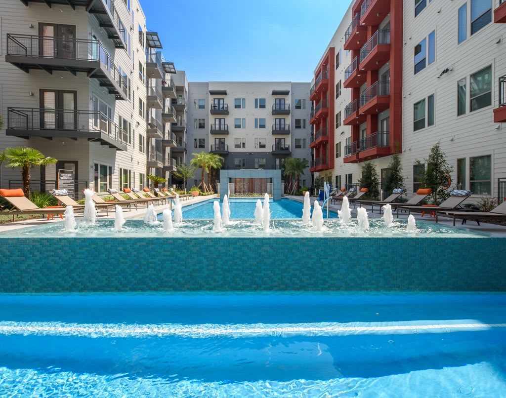 A resort-style pool area features multiple water fountains with jets, surrounded by comfortable lounge chairs and palm trees. The bright blue water reflects sunlight, and modern apartment buildings create a vibrant atmosphere in the background.