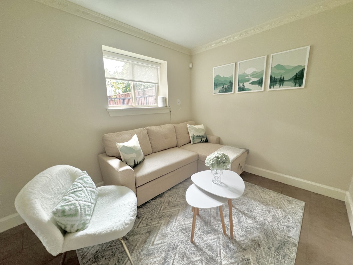 A comfortable living area features a cozy beige sofa with decorative pillows, a stylish white accent chair, and a round coffee table positioned atop a soft area rug. Framed art depicting mountain landscapes adorns the wall, while natural light enters through the window.