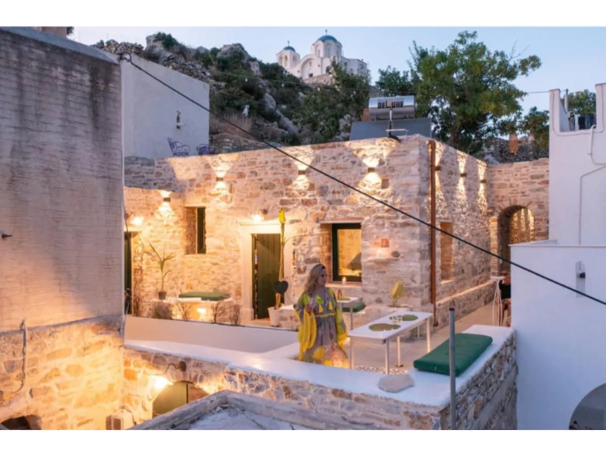 A charming outdoor dining area is visible, featuring rustic stone walls and subtle lighting. The space is adorned with green cushions and tables, inviting relaxation. In the background, the hillside showcases traditional structures, enhancing the setting's character.