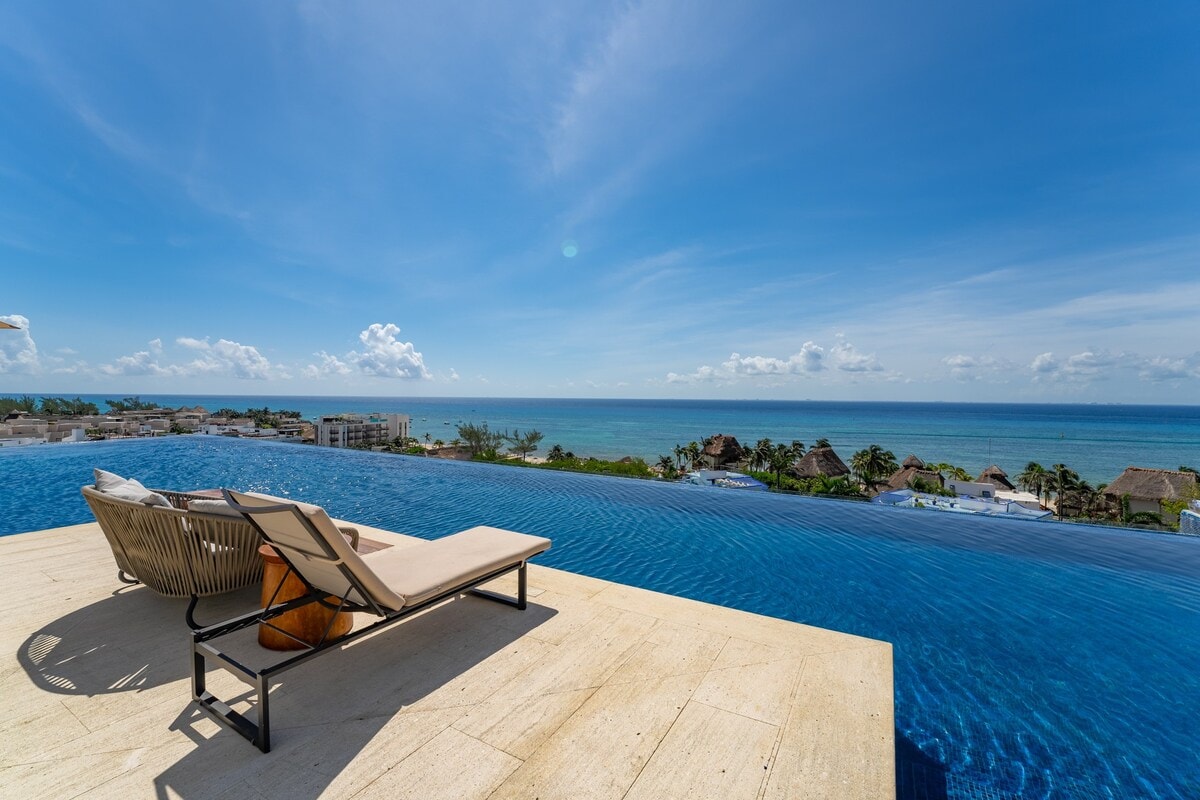 An infinity pool extends towards the horizon, reflecting the vibrant blue of the sky and ocean. A lounge chair is positioned nearby, allowing for relaxation while taking in the scenic water views. The serene atmosphere evokes a sense of tranquility.