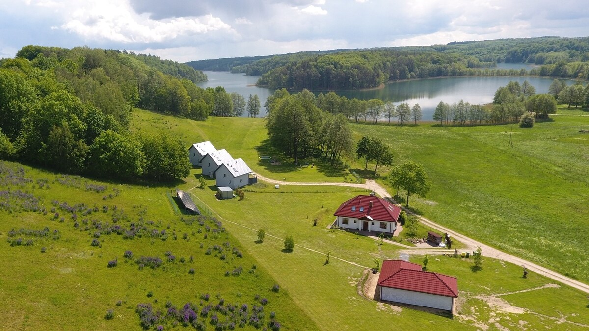 A serene landscape is depicted featuring a cluster of houses and expansive green fields, bordered by lush trees. A tranquil body of water reflects the sky, while gentle hills provide depth to the scenery. The distance showcases a natural, peaceful environment.