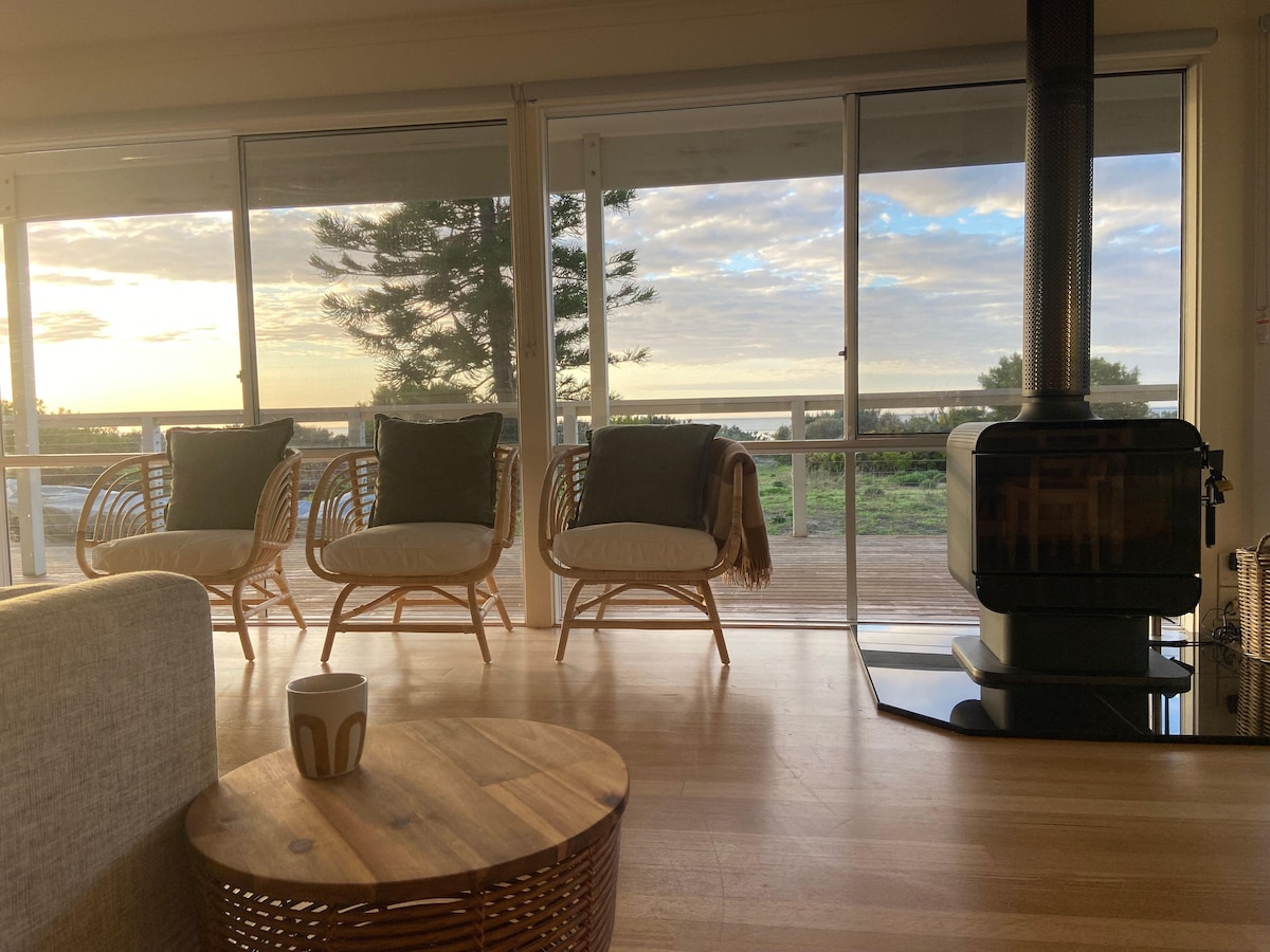 A warm living area features three single cane chairs positioned for enjoying sea views through expansive glass windows. A combustion wood heater stands elegantly in the corner, with a round wooden coffee table complementing the space's natural tones.