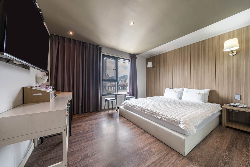 A well-lit room is furnished with a spacious bed and soft bedding. A wall-mounted television is positioned near a simple desk. Large windows are dressed with dark curtains, allowing natural light to fill the area. Wood paneling adds a touch of warmth to the space.