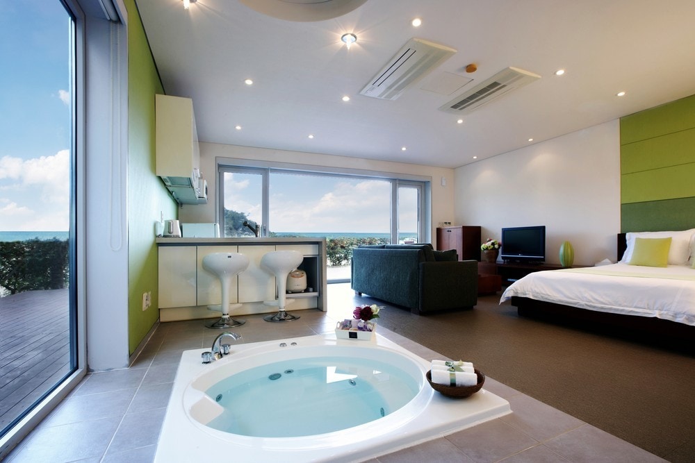 A spacious room is highlighted by a whirlpool bath and a large window offering a view of the ocean. A comfortable double bed is set against one wall, while a kitchenette and seating area are visible in the background, creating a functional layout.