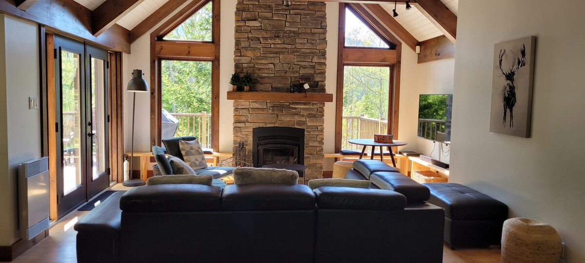 The living area features a stone fireplace as a focal point, surrounded by large windows that frame views of the greenery outside. A sectional sofa offers comfortable seating, and a coffee table sits nearby. Light floods the space, accentuated by the wooden beams overhead.