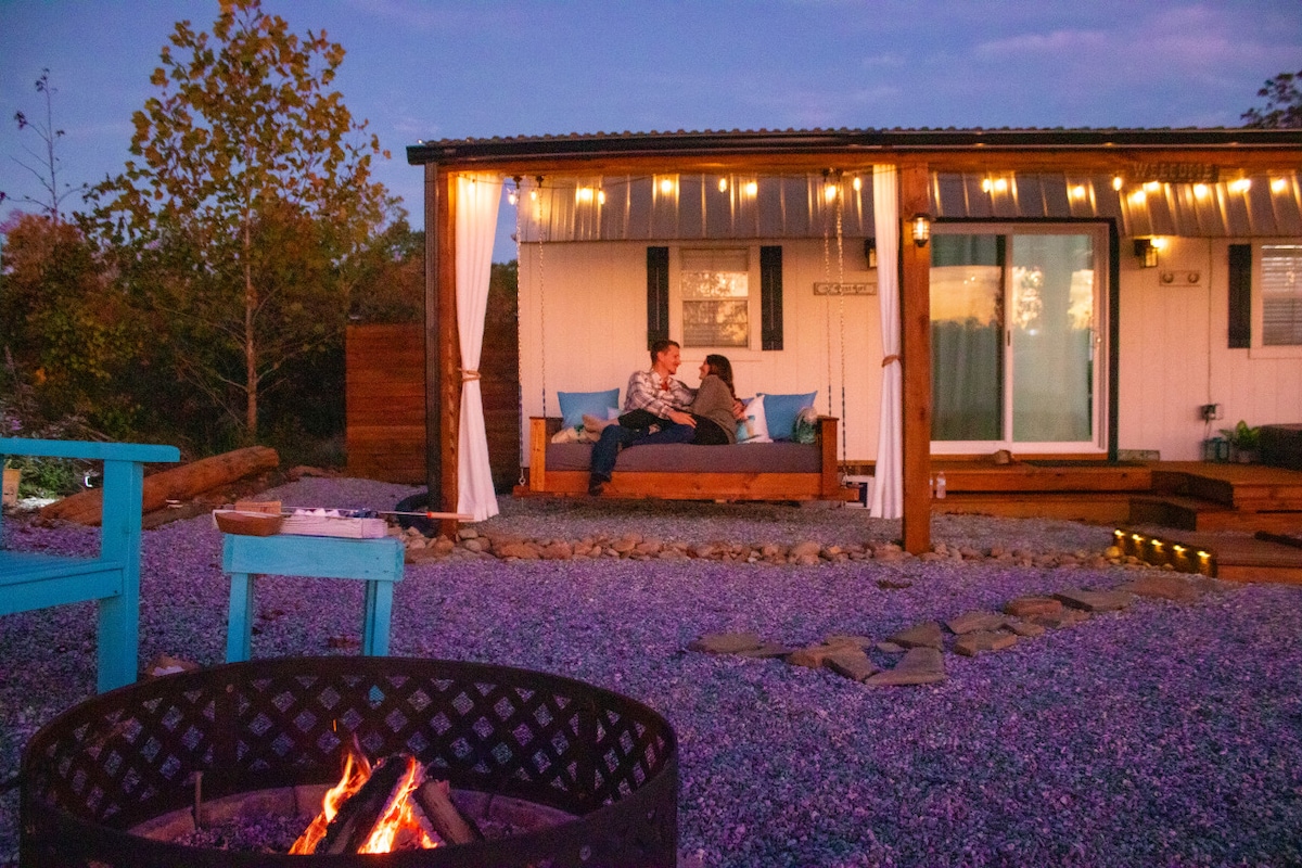 An outdoor seating area is illuminated by string lights, featuring a cozy porch bed swing adorned with plush cushions. A fire pit with a controlled flame serves as a focal point, surrounded by seating, while the exterior of the cottage is framed by soft landscaping.
