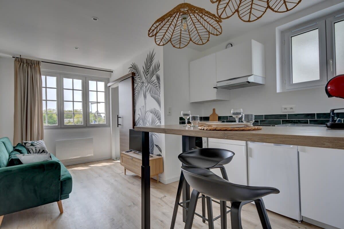A modern kitchen and living area is displayed, featuring a compact dining table with high black chairs. A green sofa is positioned against a light gray wall adorned with decorative plants. Natural light floods in through large windows, creating a bright atmosphere.