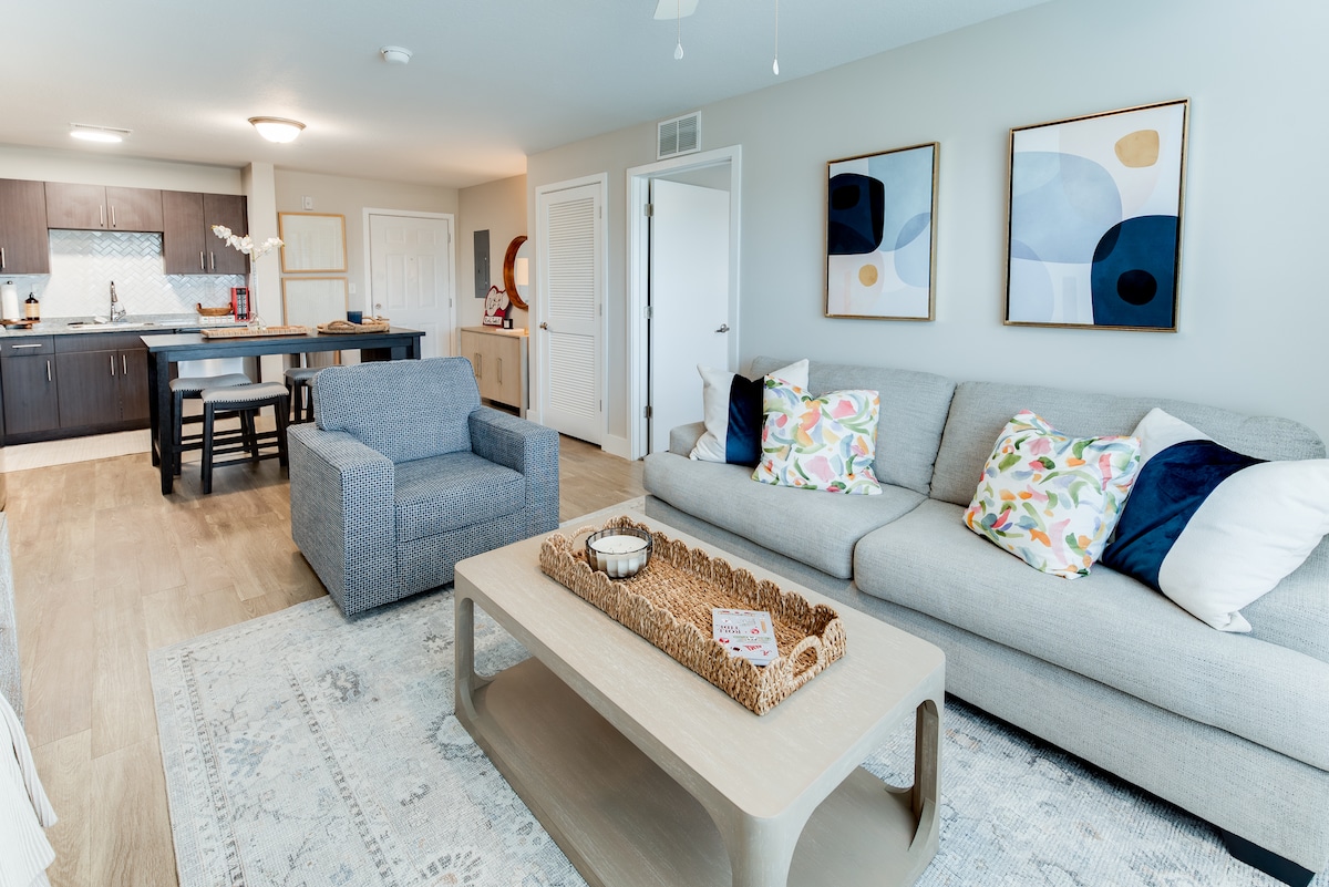 An open-concept living area features a light gray sofa adorned with colorful cushions, complemented by a textured coffee table. A cozy armchair is situated nearby, and a dining table is visible in the background, adjacent to a modern kitchen with dark cabinetry.