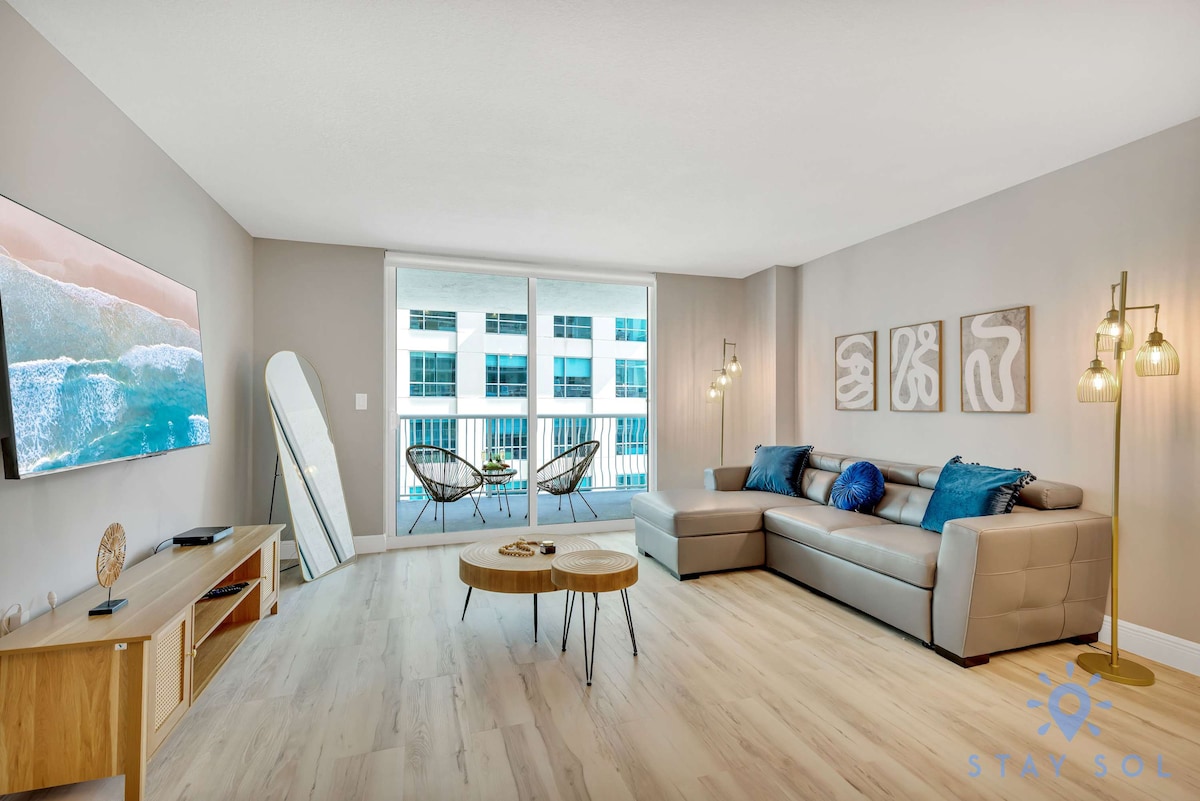 A modern living area is showcased, featuring a sectional sofa paired with vibrant blue pillows. A wooden coffee table sits in front of the sofa, complemented by stylish floor lamps. Large windows allow natural light to fill the space and offer views of the outdoor balcony.