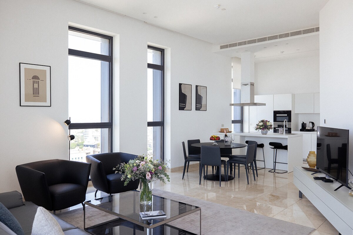 A modern living space features large windows allowing natural light to fill the room. Chic black armchairs complement a sleek coffee table. A dining area with stylish chairs is visible, alongside a contemporary kitchen outfitted with minimalistic cabinetry and appliances.