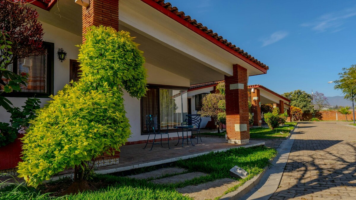 The image features a row of charming villas with terracotta roofs, set against a clear blue sky. Lush greenery and manicured shrubs frame the entrance, while a small outdoor seating area with chairs is visible on a paved pathway leading to each villa.