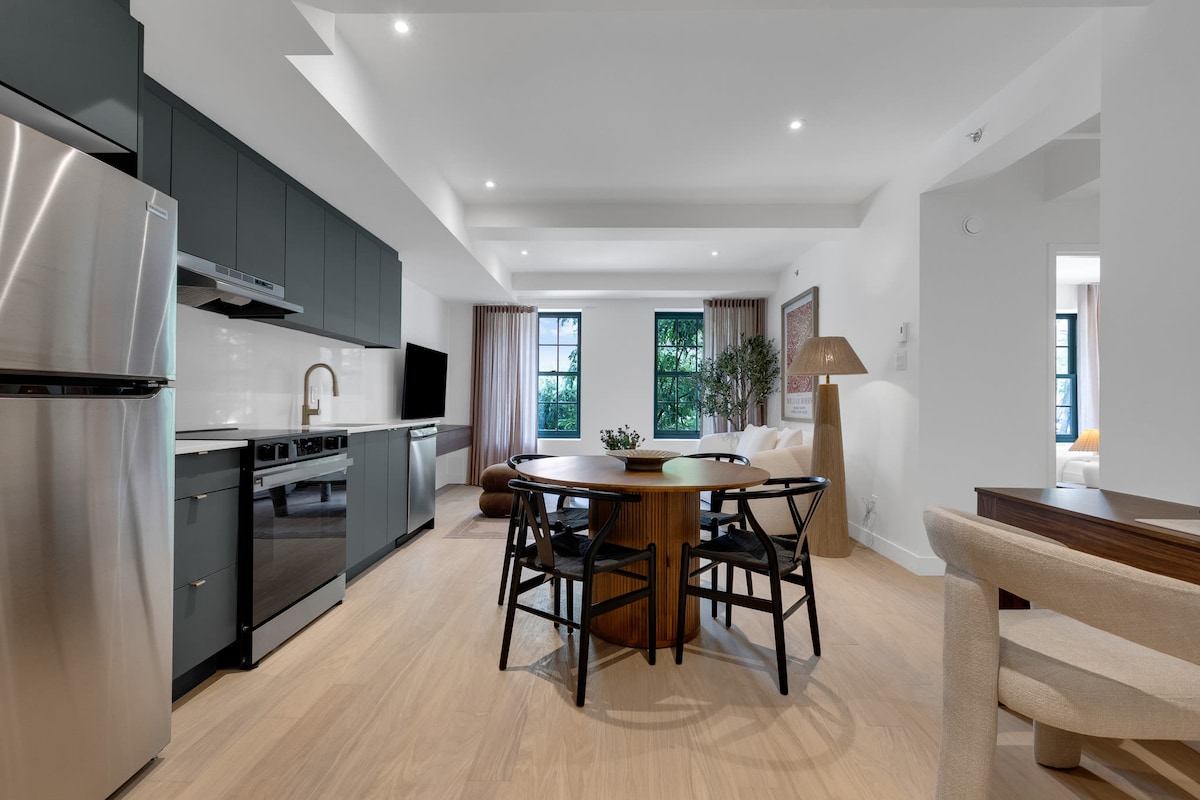 A well-lit open-concept living area features a modern kitchen alongside a spacious dining table. The sleek cabinetry complements stainless steel appliances. Large windows allow natural light to fill the space, enhancing the inviting atmosphere of the room.