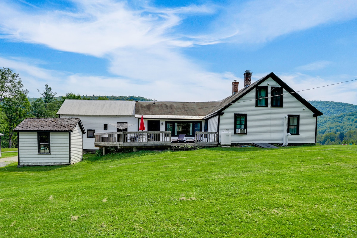A spacious home is featured, set against a backdrop of rolling green hills. The exterior showcases a large deck with seating areas and an umbrella. A small storage shed is positioned nearby, completing the inviting outdoor landscape.
