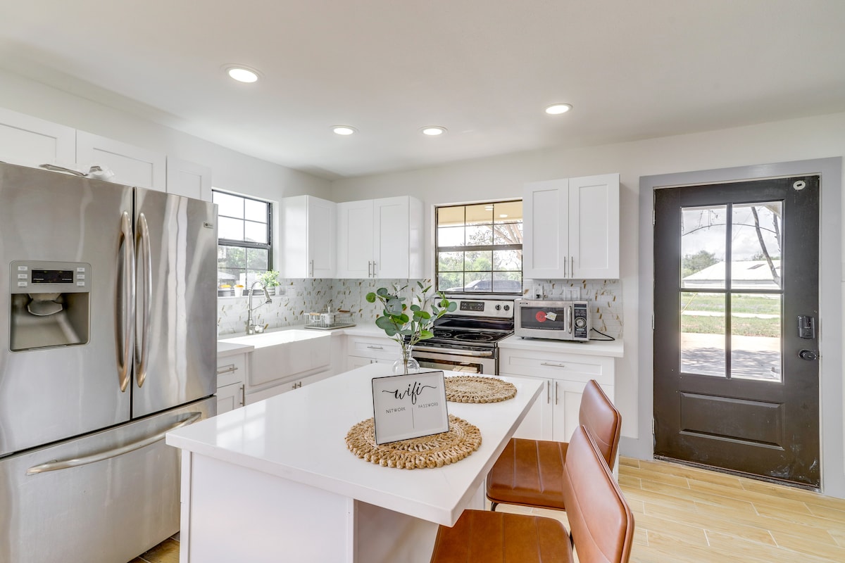 A modern kitchen features white cabinetry and sleek stainless-steel appliances. The island with seating is accompanied by a decorative sign. Abundant natural light fills the space through multiple windows, enhancing the clean and functional design.
