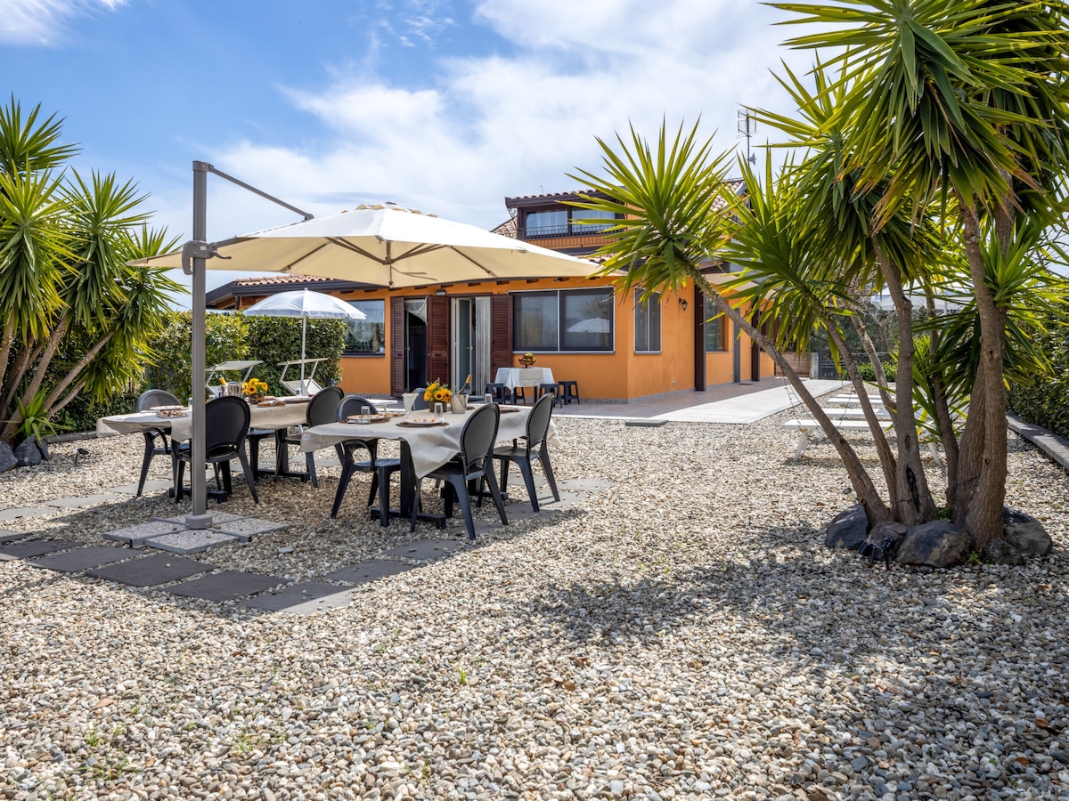 An outdoor dining area features several tables and chairs under large umbrellas, surrounded by landscaped gravel and palm trees. In the background, the property displays a warm exterior with windows and a patio space, set against a clear blue sky.