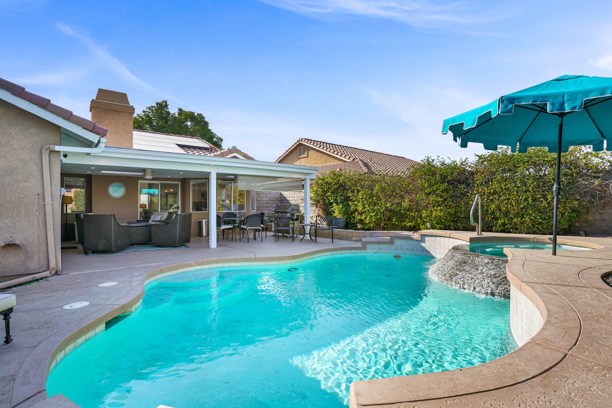A heated pool with a soothing waterfall feature is surrounded by a spacious patio area, offering ample seating. An umbrella provides shade for relaxation. The exterior of the home, featuring a glass sliding door, complements the inviting atmosphere of the outdoor space.
