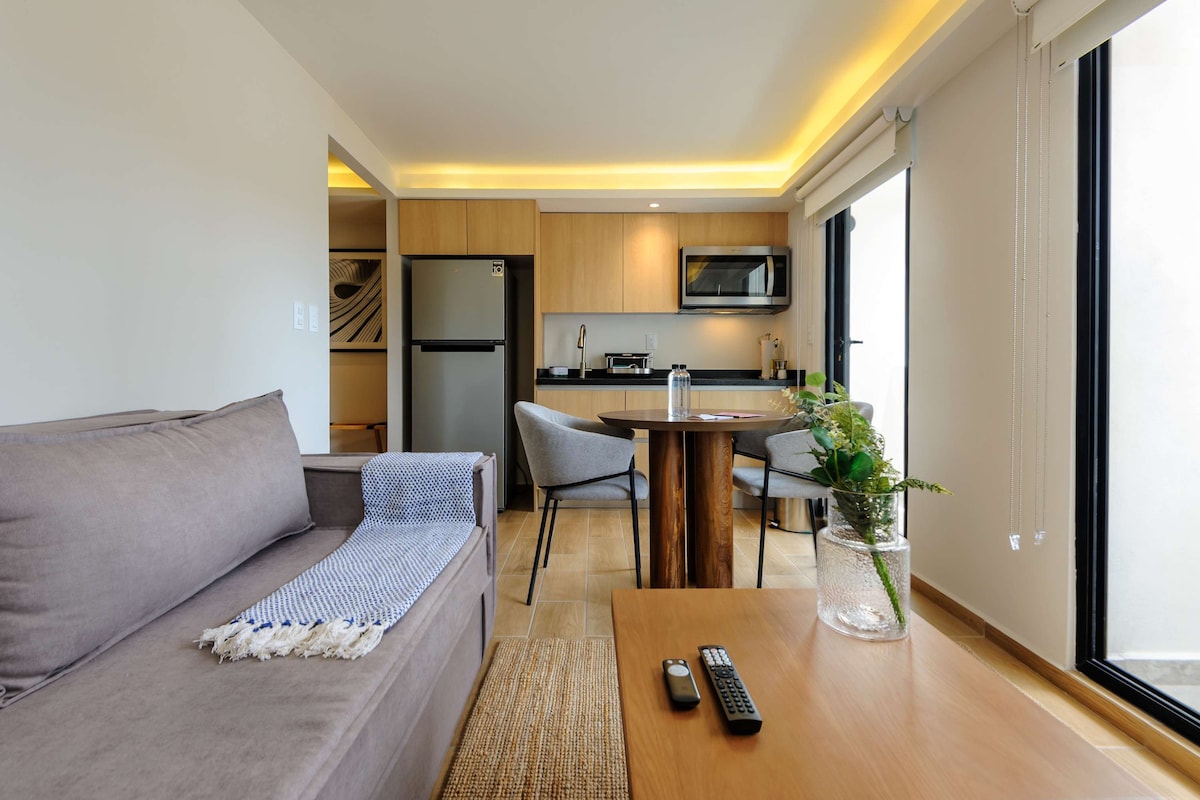 A bright living area features a comfortable couch and a wooden dining table surrounded by two chairs. Modern kitchen appliances are visible in the background, along with warm lighting that enhances the inviting atmosphere. Large glass doors open to a small balcony, allowing for natural light.