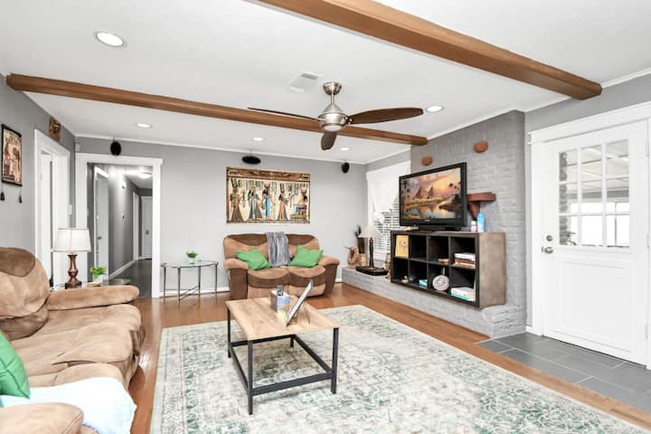 Gezellige woonkamer met twee bruine banken, groene kussens en een bijpassend tapijt. Beschikt over een grote tv, een bruine salontafel en drie glazen bijzettafels. Ingericht met elegante oude Egyptische kunst voor een warme, stijlvolle en cultureel rijke sfeer.