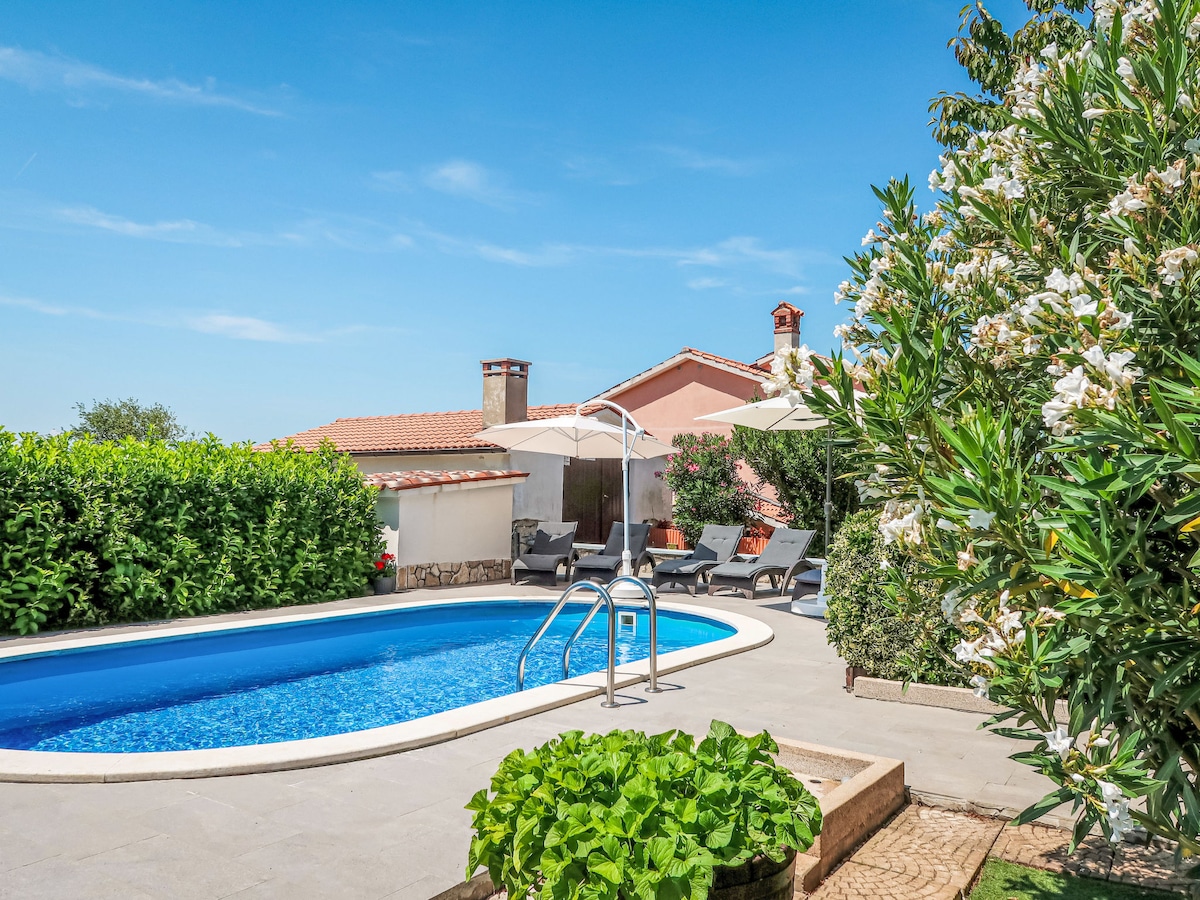 A round swimming pool is featured, surrounded by sun loungers and umbrellas. Lush greenery and blooming flowers add a vibrant touch to the outdoor space, creating a serene atmosphere. The house exterior is visible in the background, providing a picturesque setting.