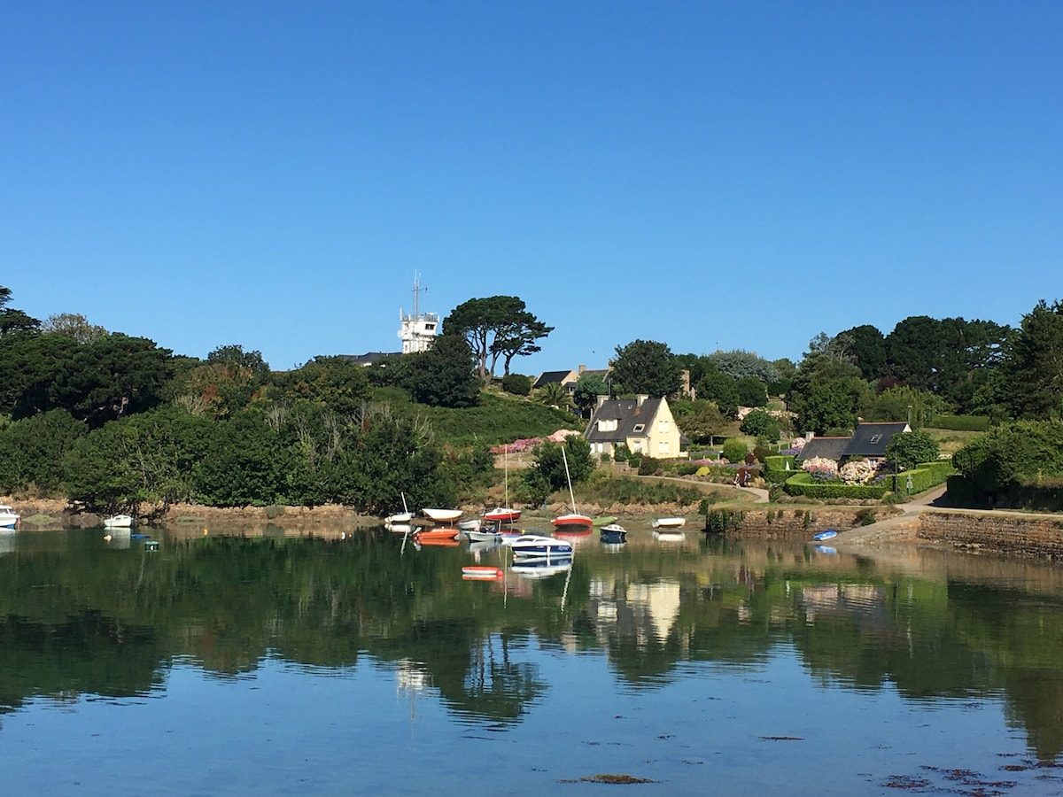 A serene coastal scene illustrates a charming house set against lush greenery, reflecting on calm waters. Several boats are anchored nearby, while a lighthouse is visible on the horizon, contributing to the peaceful ambiance of the Île de Bréhat.