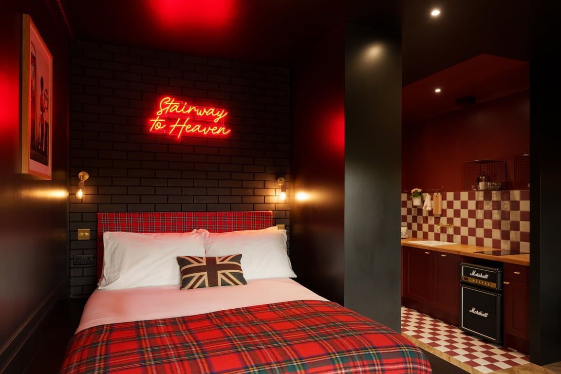 A vibrant studio features a comfortable double bed draped in a tartan blanket, with a striking neon sign above stating 'Stairway to Heaven.' The kitchenette is visible in the background, showcasing bold red and white checkered tiles along with modern appliances.
