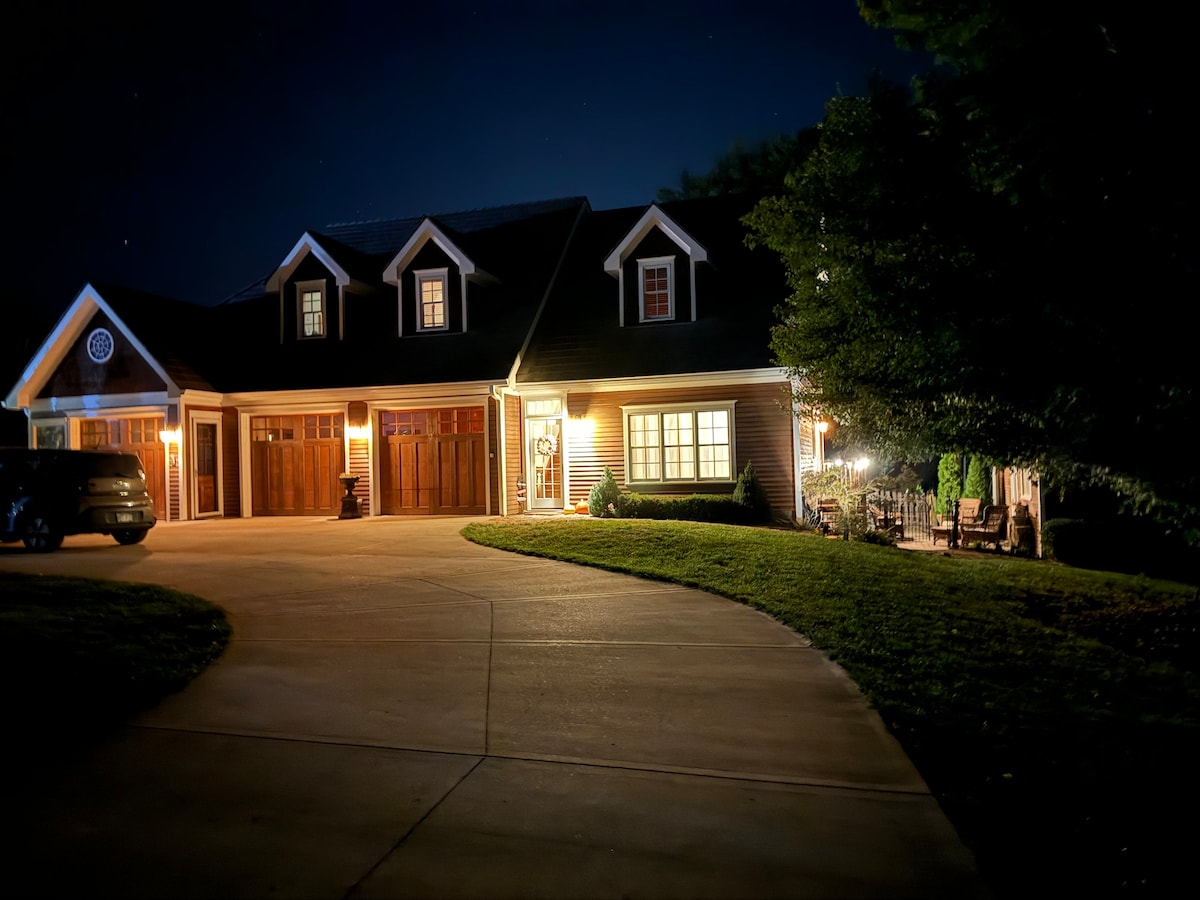 A well-lit exterior of a multi-gabled house is visible at night, featuring wooden garage doors and a welcoming front entrance. A circular driveway encircles a lawn, and the outdoor seating area is subtly illuminated, creating a serene evening ambiance.