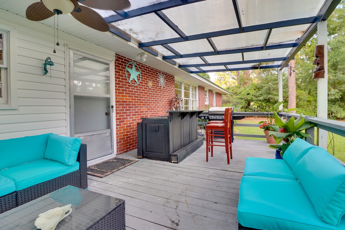 A covered patio features teal seating and a dining area with bar stools, complemented by a gas grill and outdoor bar. The area is surrounded by greenery, providing a serene outdoor space for relaxation and dining.