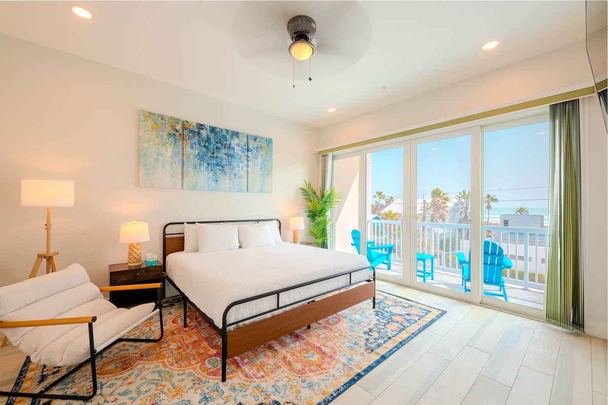 A spacious bedroom features a comfortable bed with a modern metal frame, complemented by a stylish area rug. Large glass doors lead to a balcony with seating, allowing natural light to fill the room. An art piece adds a touch of color to the serene atmosphere.