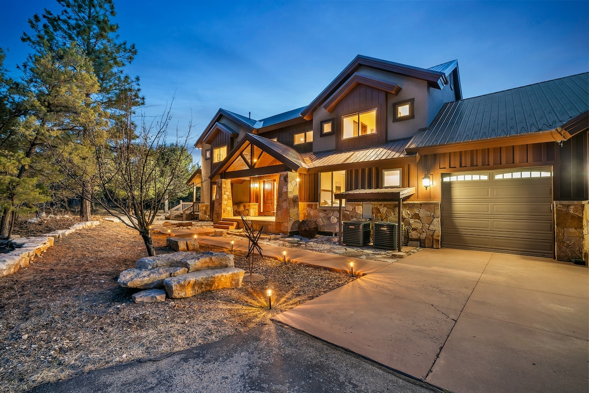 A welcoming exterior of a mountain retreat is presented, showcasing a blend of stone and wood materials. The well-lit pathway leads to the entrance, framed by soft landscaping and tall pine trees, under a clear evening sky.