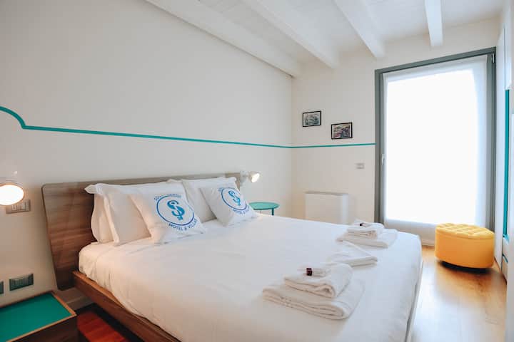 Habitación doble luminosa con muebles modernos y tonos naturales, con grandes ventanales que garantizan luz y comodidad para una estancia relajante en Peschiera del Garda.